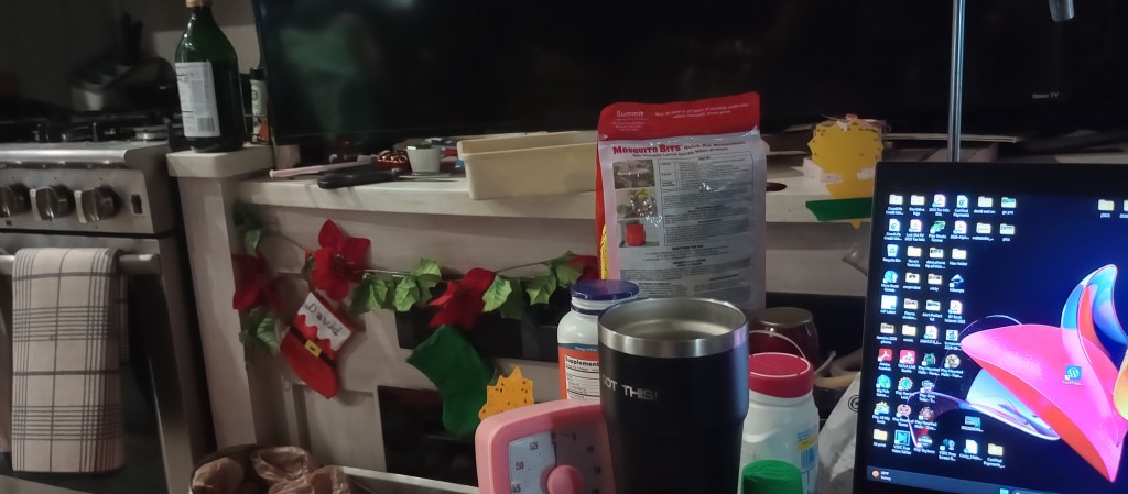 A cluttered kitchen countertop featuring a laptop, various kitchen supplies, a coffee mug, and festive decorations including stockings.